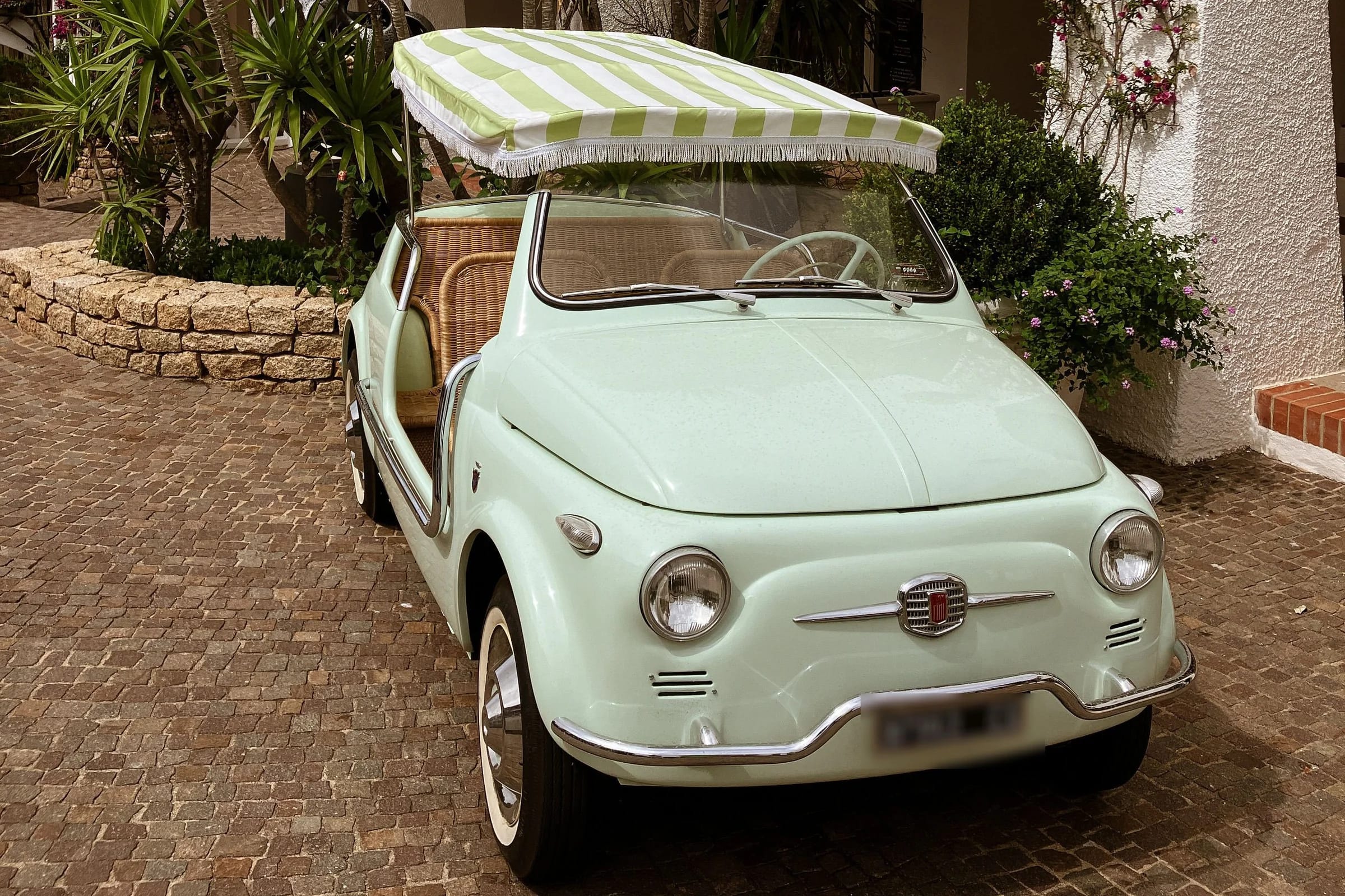 A mint-green open-topped Fiat 500 with a striped canopy and wicker seats, parked on cobblestones before a whitewashed wall